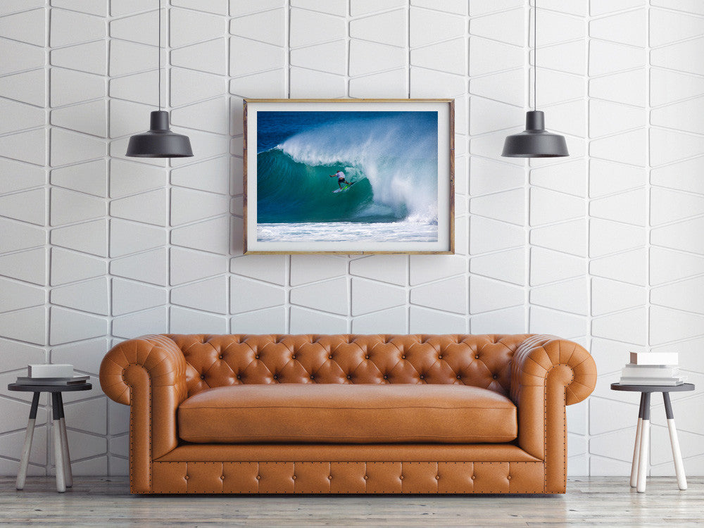 The Mick Fanning barrel wave print shown framed in light oak wood, above a tan leather sofa.