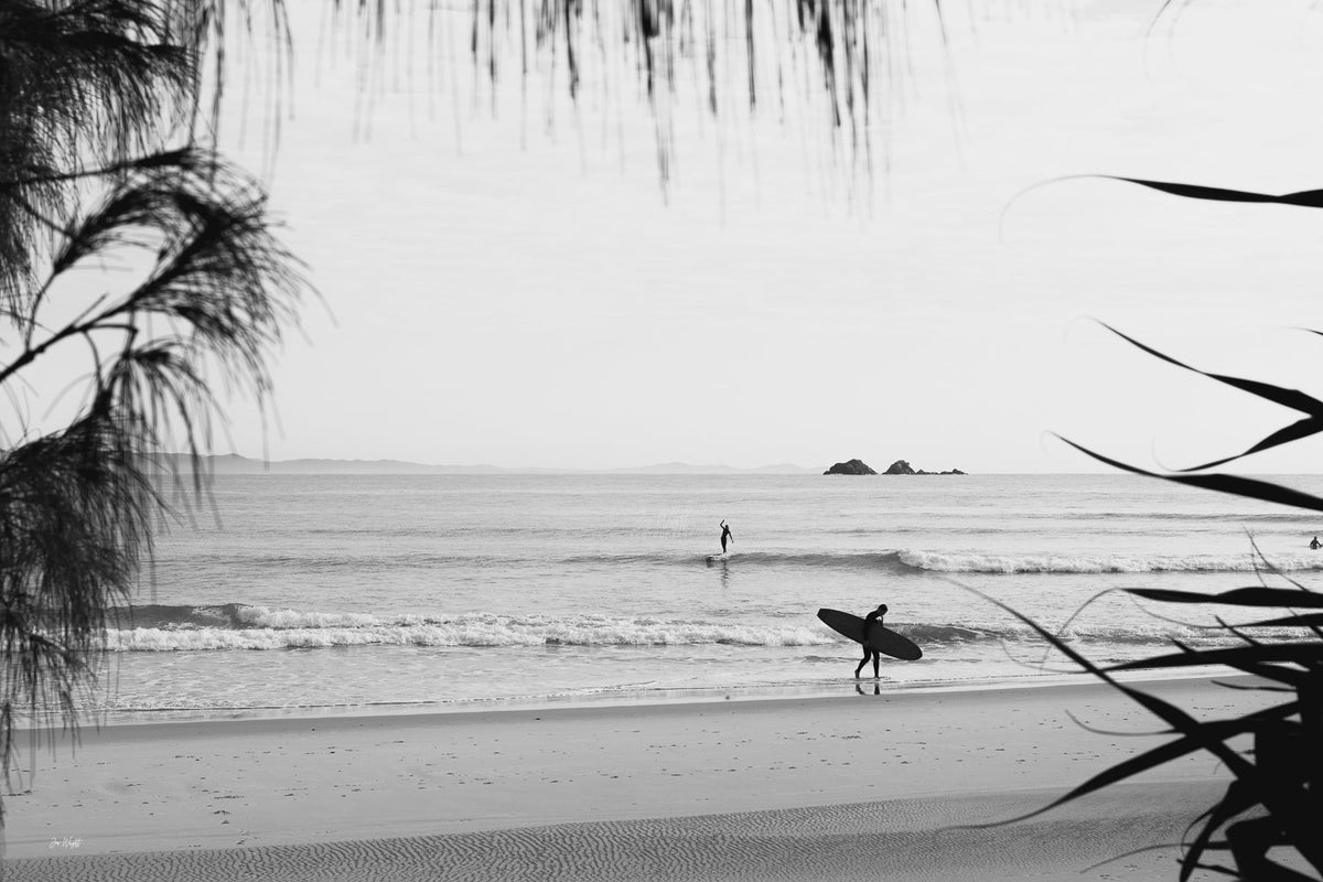 Black and white morning at Byron Bay, a longboarder walking the wet sand under casuarina needles, a surf wall print.
