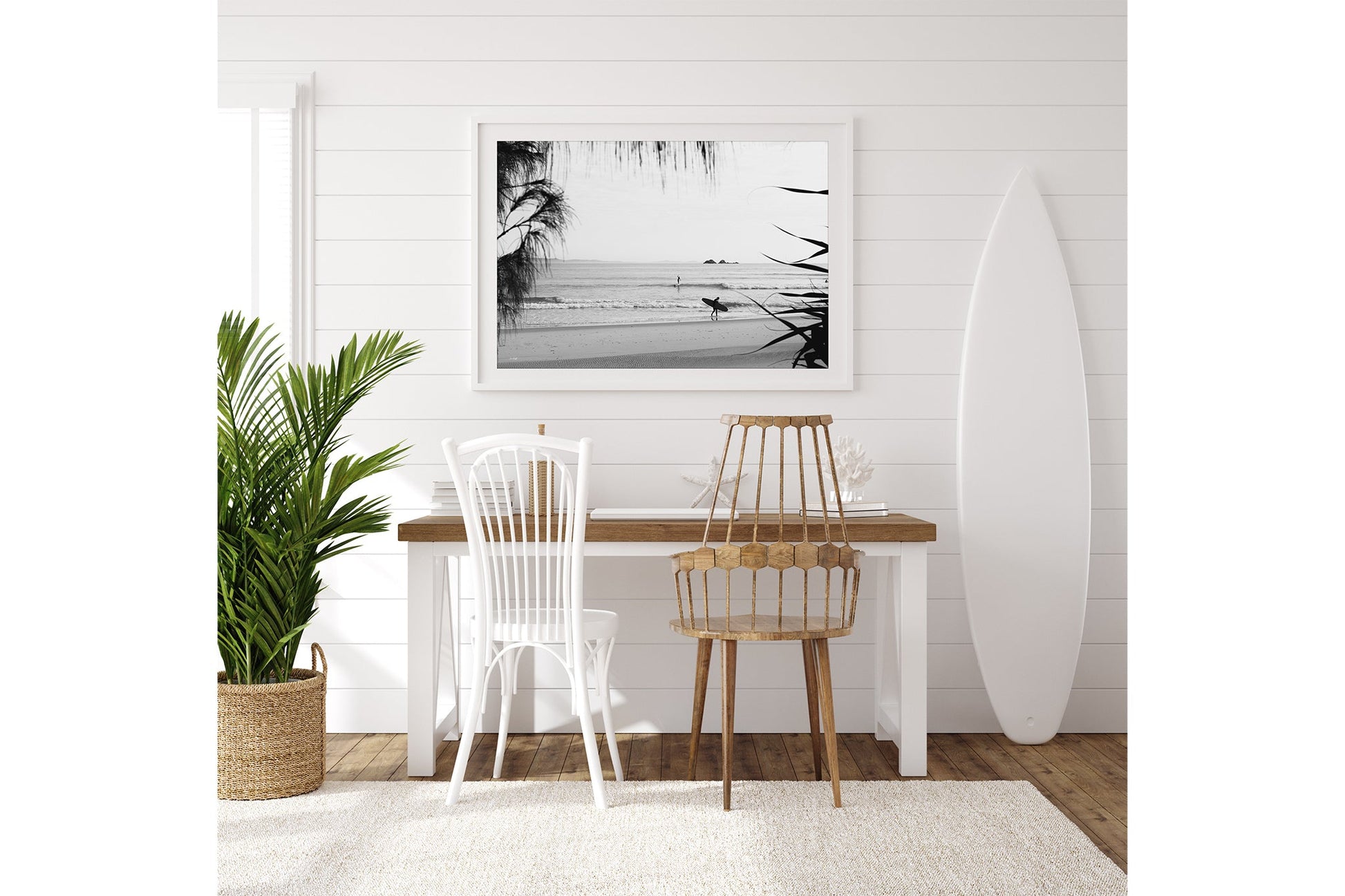 Byron Bay black and white surf print shown framed in white beside a surfboard in a coastal room.
