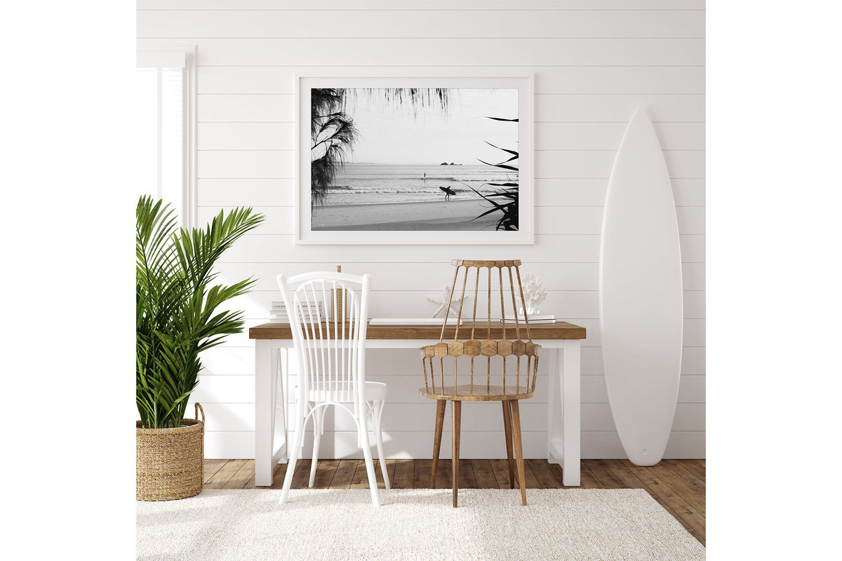 Byron Bay black and white surf print shown framed in white beside a surfboard in a coastal room.