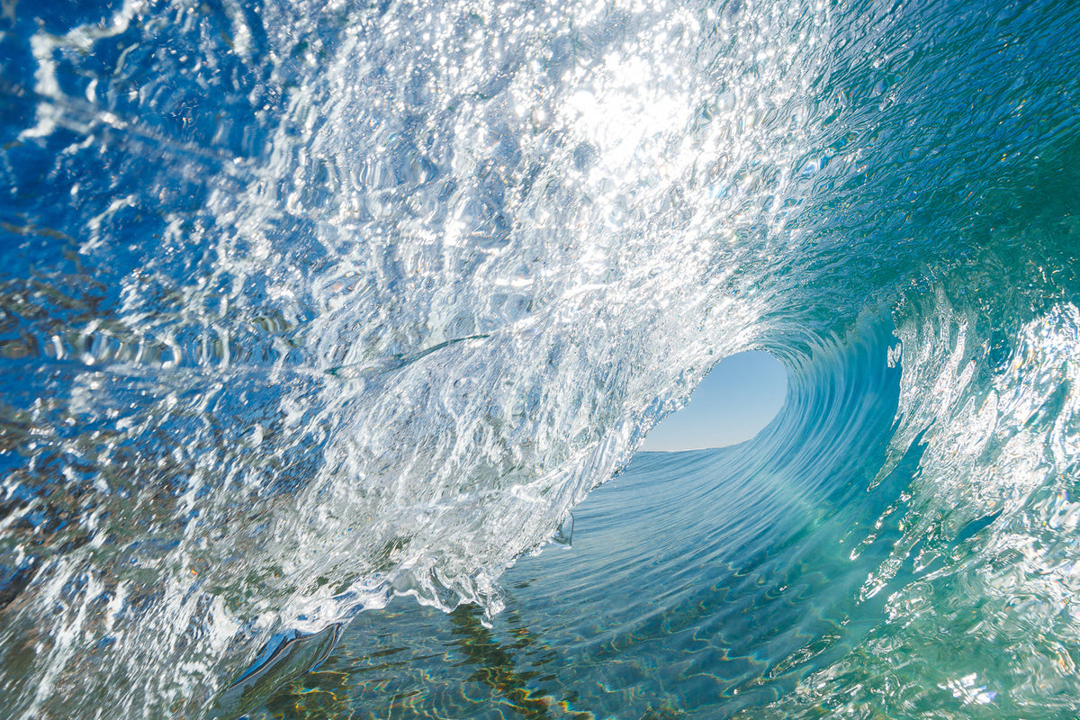 Inside a clear barrelling wave, sunlit white spray on the left and aqua water curling toward a patch of pale sky, an ocean wall print.