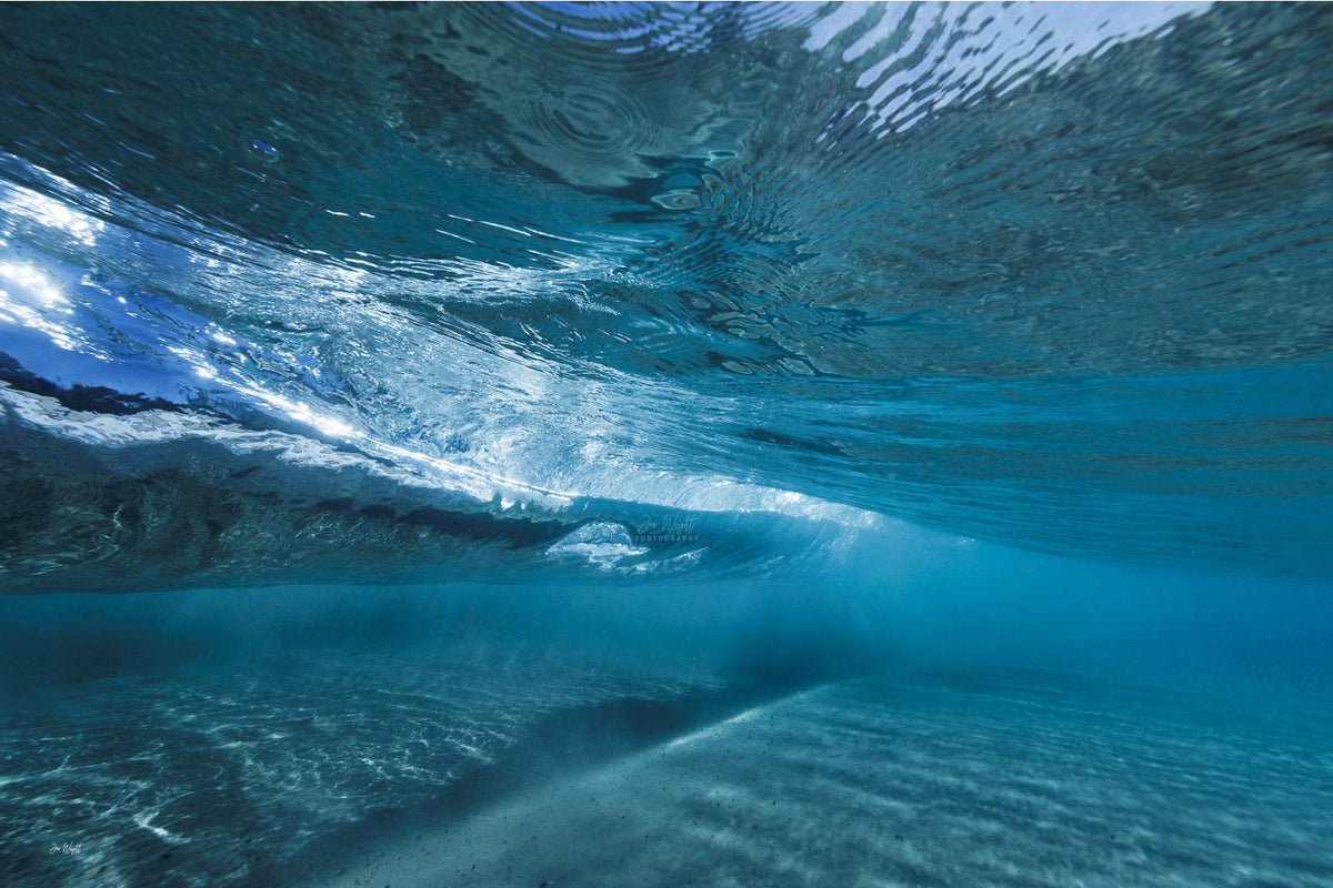 Underwater view of a wave passing overhead, rippled teal and blue water above a sandy seabed with soft light, an underwater wall print.