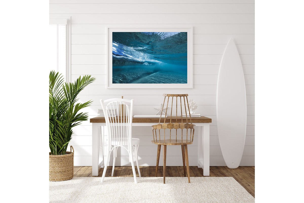 Quiet Shallows print shown framed in white on a white shiplap wall with a surfboard leaning beside a desk.