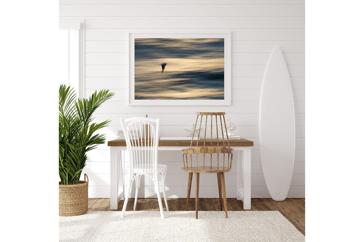 Gliding Gull print shown framed in white on a white shiplap wall with a surfboard leaning beside a desk.