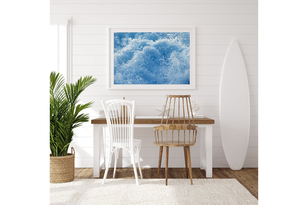 Cobalt Chaos print shown framed in white on a white shiplap wall with a surfboard leaning beside a desk.
