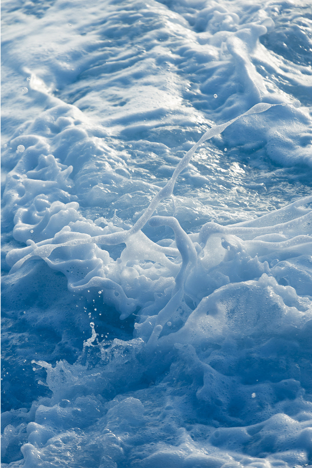 A thin white ribbon of foam arcing across layers of blue and white sea froth with fine droplets, an abstract ocean wall print.