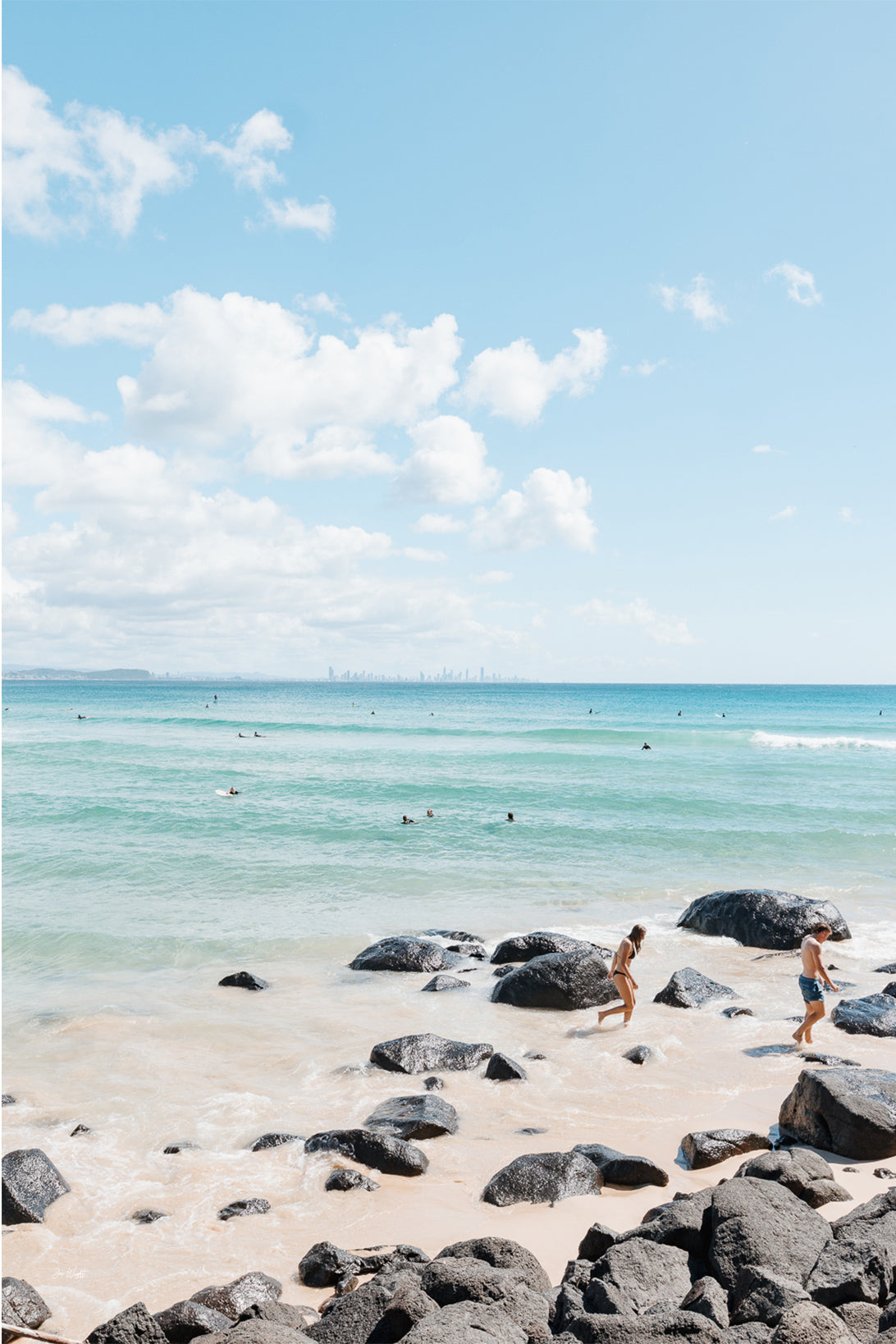 Dark boulders on pale sand with two people walking beside turquoise water and a faint skyline on the horizon, Gold Coast, a beach print.