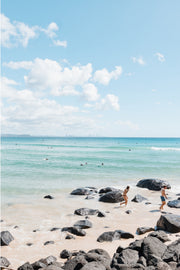 Dark boulders on pale sand with two people walking beside turquoise water and a faint skyline on the horizon, Gold Coast, a beach print.
