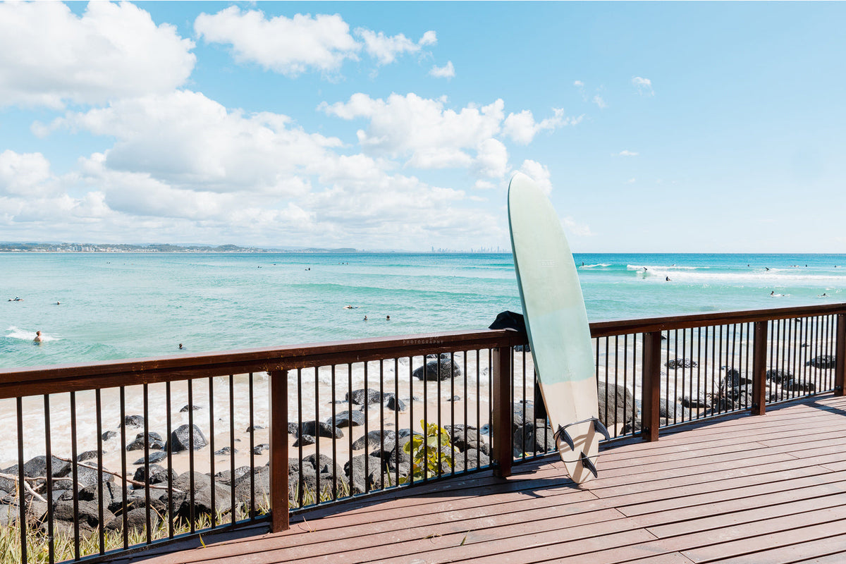 A pale surfboard leaning on a timber boardwalk rail above rocks, turquoise water and surfers, Gold Coast, a beach wall print.