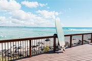 A pale surfboard leaning on a timber boardwalk rail above rocks, turquoise water and surfers, Gold Coast, a beach wall print.