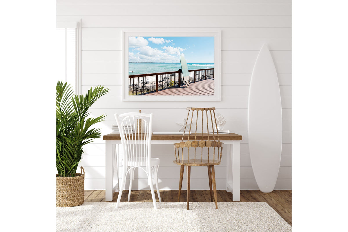 Boardwalk Break print shown framed in white on a white shiplap wall with a surfboard leaning beside a desk.