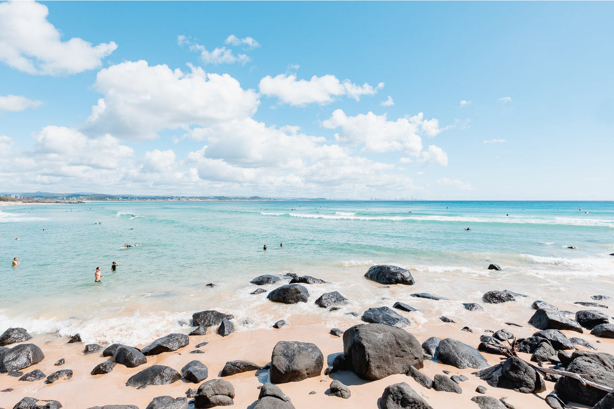 Dark boulders on pale sand beside turquoise shallows with swimmers and big white clouds, Gold Coast, a beach wall print.