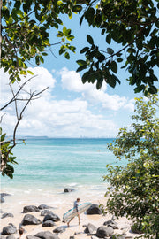 A surfer carrying a white board across pale sand, framed by leaves above turquoise water and a faint skyline, Gold Coast, a beach print.