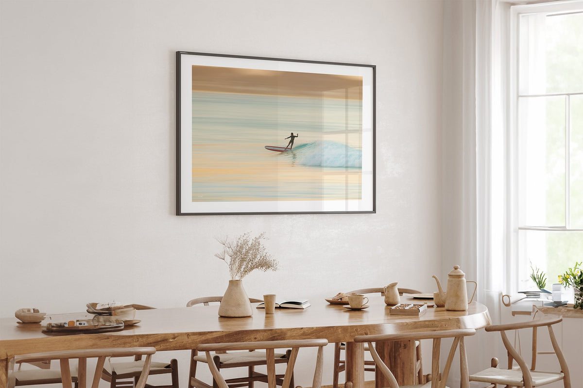 Longboard surfer print shown framed in black above an oak table.