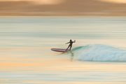 A longboard surfer in a black wetsuit on a red and white board drops into a small turquoise wave, with soft peach, gold and aqua motion blur behind, a surf wall print.