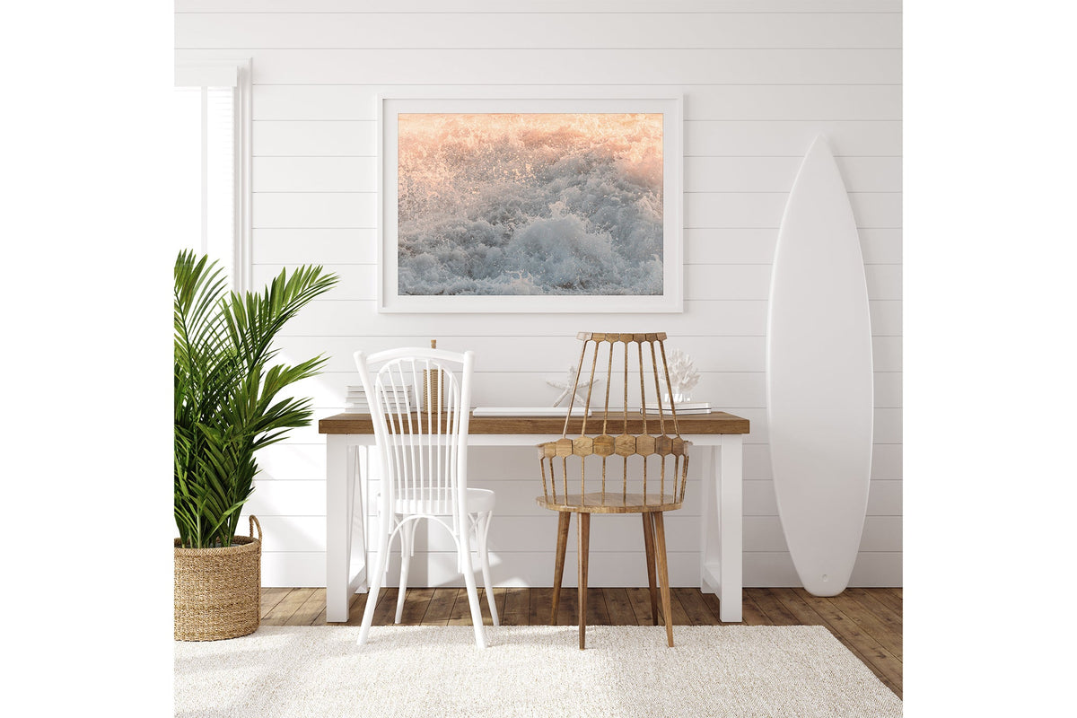 Peach Spray print shown framed in white on a white shiplap wall with a surfboard leaning beside a desk.