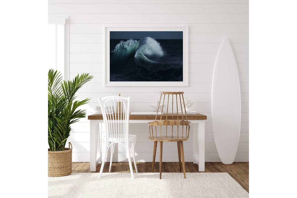 Moonlit Curl print shown framed in white on a white shiplap wall with a surfboard leaning beside a desk.