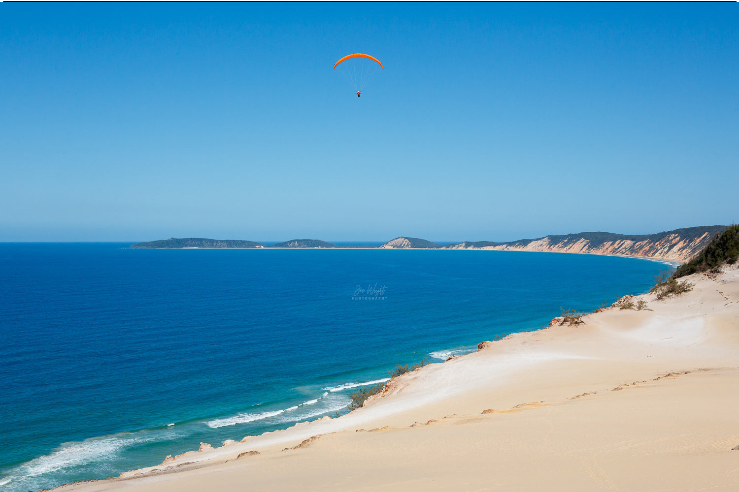 An orange paraglider hangs above deep blue ocean and the coloured sand cliffs of Rainbow Beach, a coastal wall print.