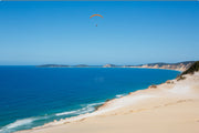 An orange paraglider hangs above deep blue ocean and the coloured sand cliffs of Rainbow Beach, a coastal wall print.