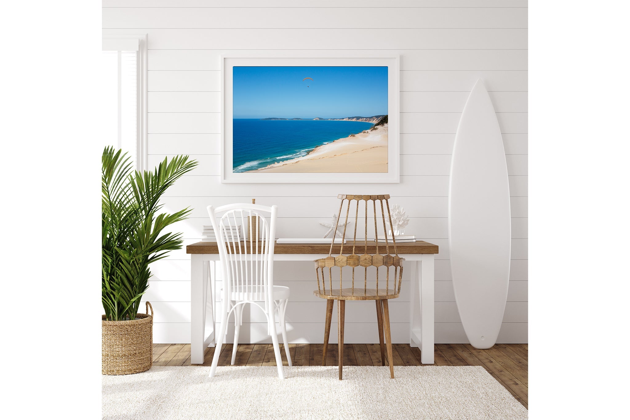 Rainbow Beach paraglider print shown framed in white above a desk beside a surfboard.