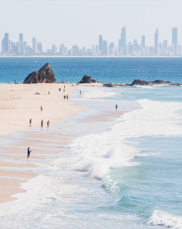 Currumbin Beach's rock outcrop and shoreline with walkers on the sand, Surfers Paradise skyline hazy beyond, a print.
