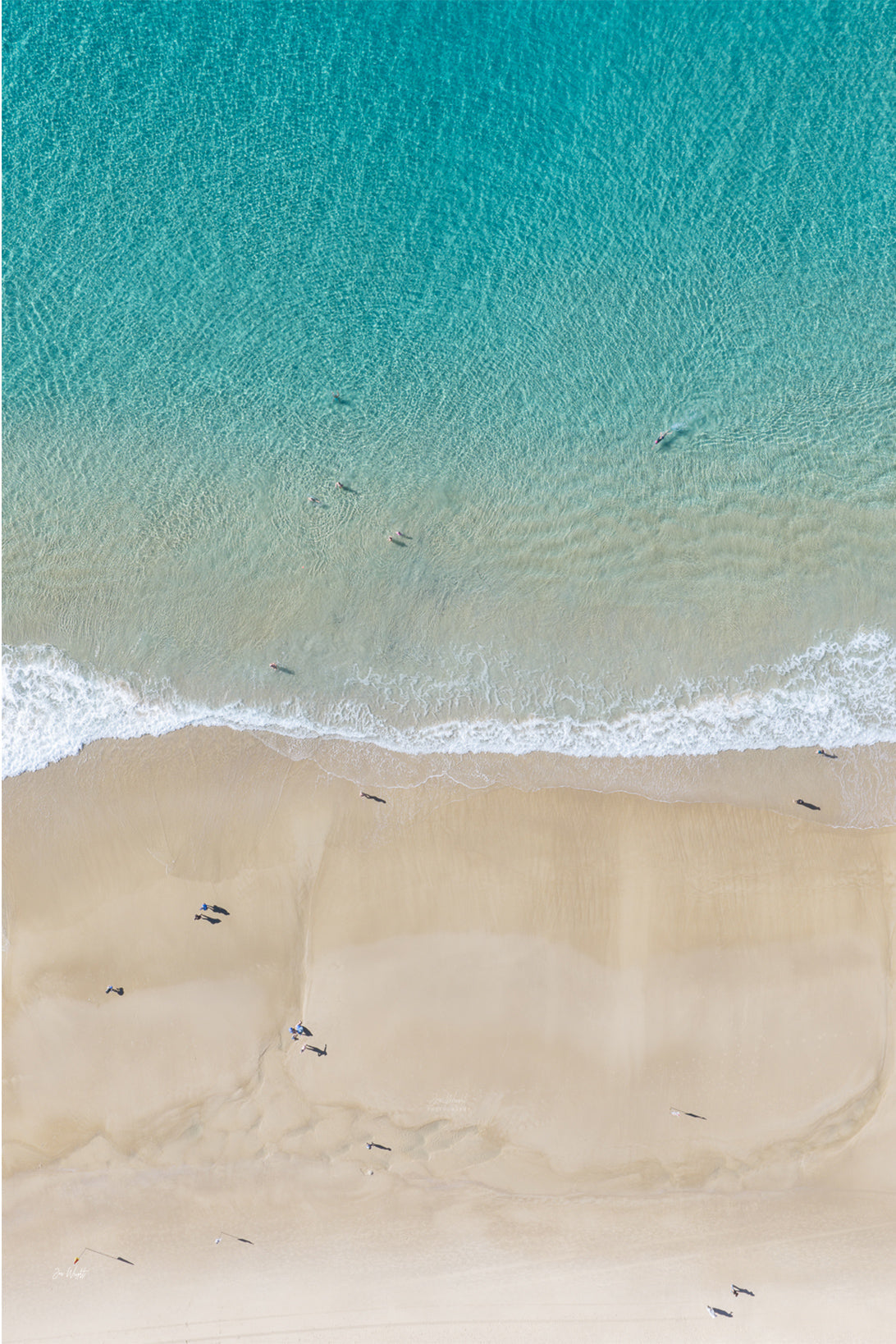 Aerial view of rippled turquoise shallows meeting pale sand, with a few swimmers scattered through the water.