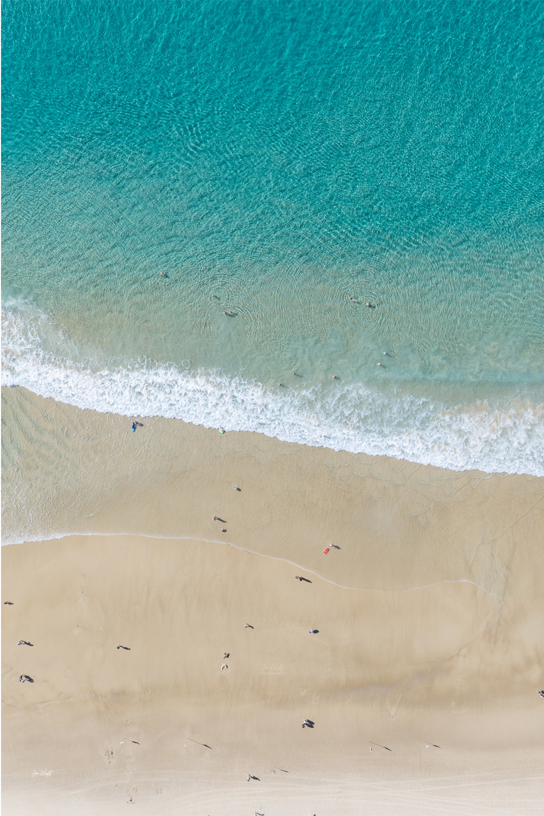 Aerial view of turquoise water breaking onto pale sand, dotted with sunbathers and swimmers along the shoreline.