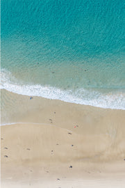 Aerial view of turquoise water breaking onto pale sand, dotted with sunbathers and swimmers along the shoreline.