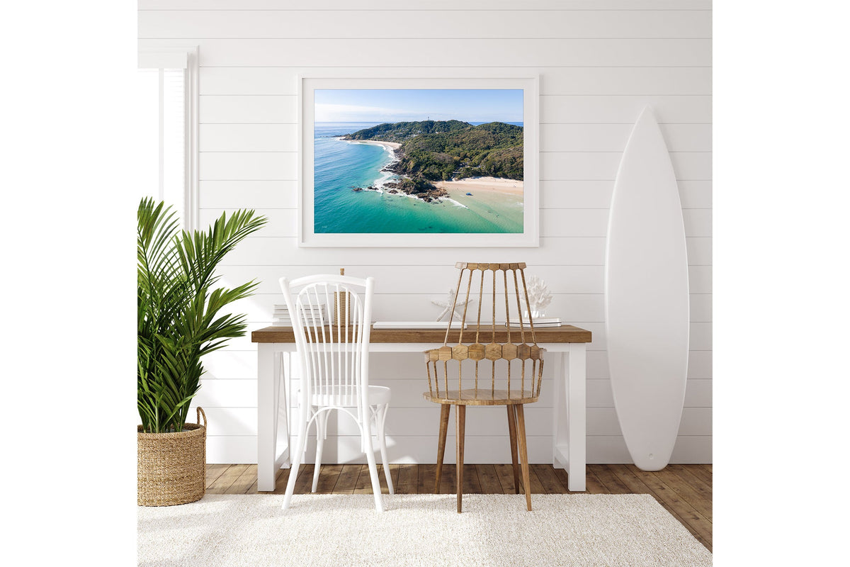 Cape Byron aerial print shown framed in white beside a surfboard in a coastal room.