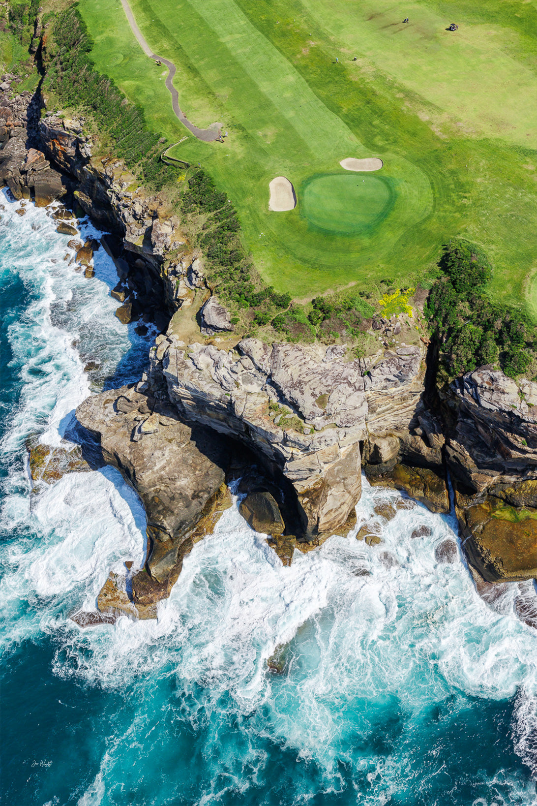 Golf course with green fairway and sand bunkers, surrounded by rocky cliffs and crashing ocean waves.
