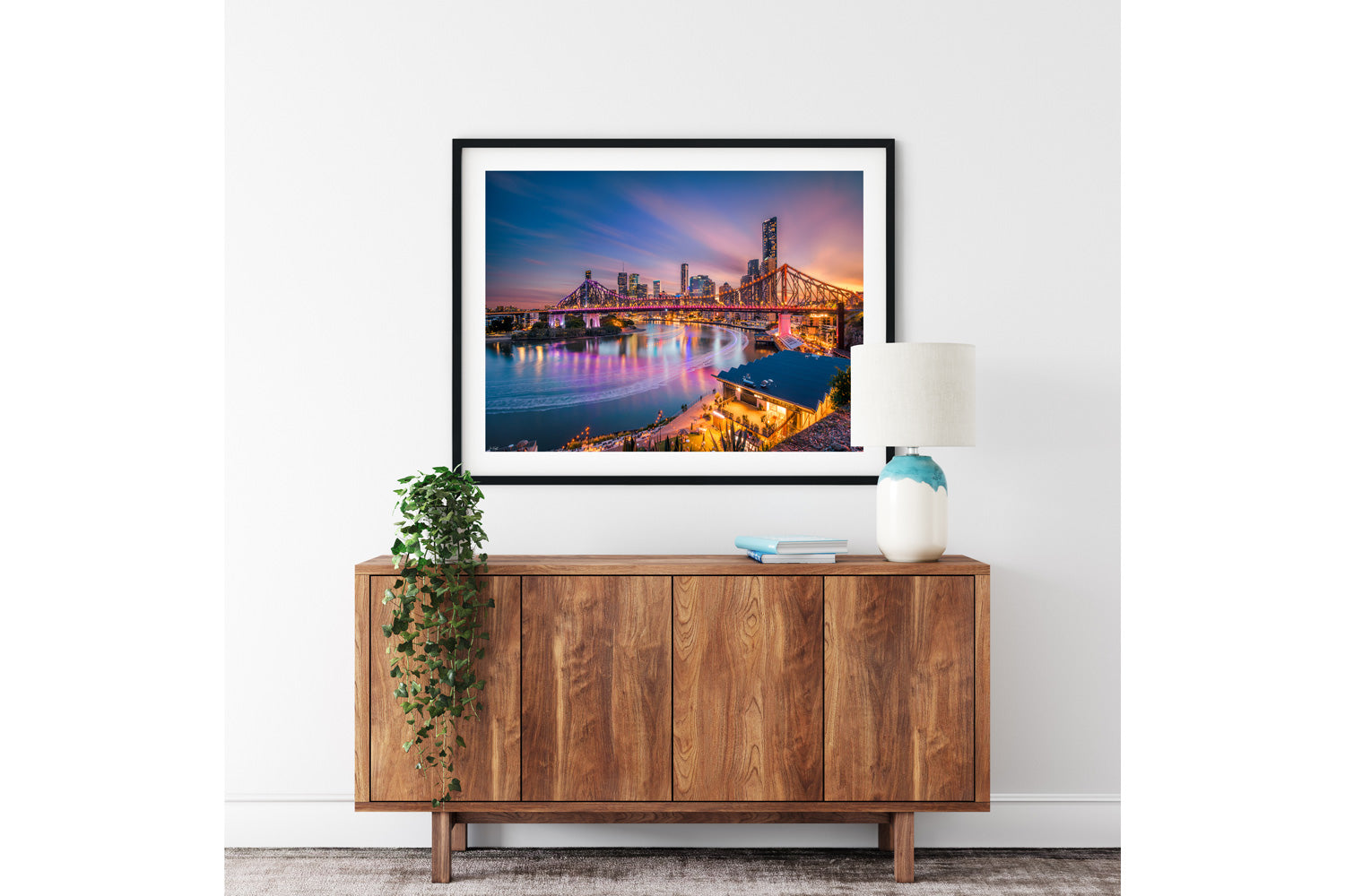 The Story Bridge dusk print shown framed in black, above a wooden sideboard with a lamp.