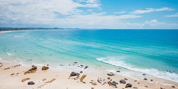 Aerial view along Kirra Beach, pale turquoise shallows meeting a sandy shoreline with scattered rocks, Gold Coast skyline in the distance.